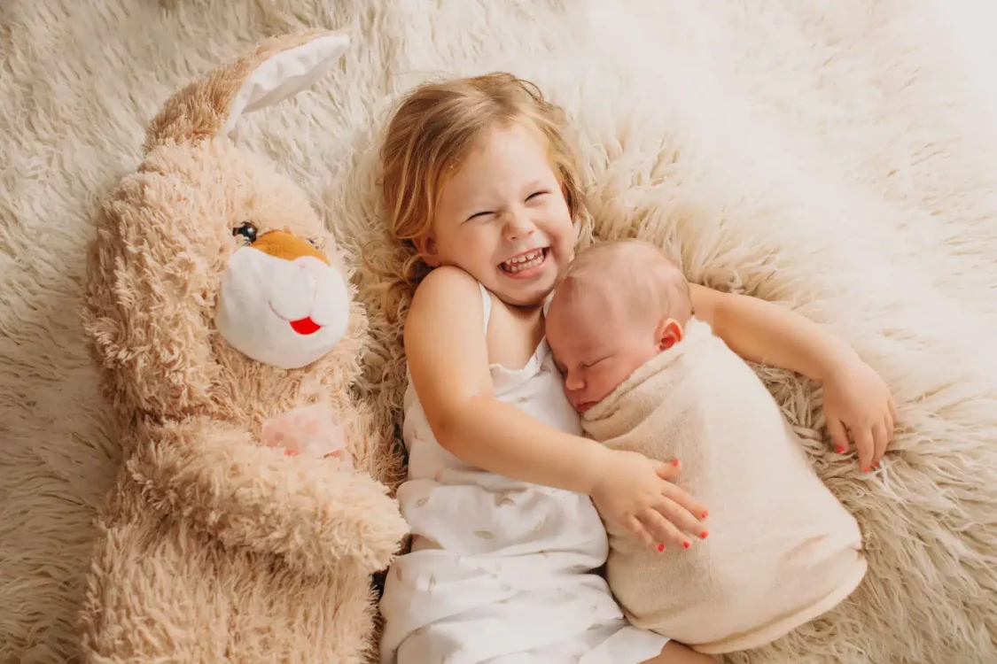 Regard pétillant d'une grande sœur tenant dans ses bras son petit frère nouveau-né, studio photo Brabant Wallon